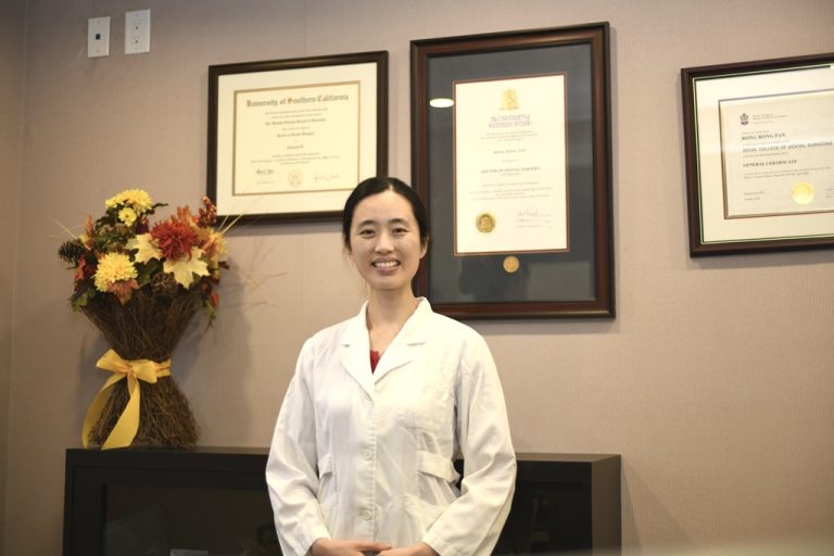 Dr. Cindy Li standing and smiling in a white coat in front of framed certificates at Excel Dental.