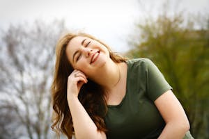 Smiling young woman outdoors with her head tilted and hand resting near her face.