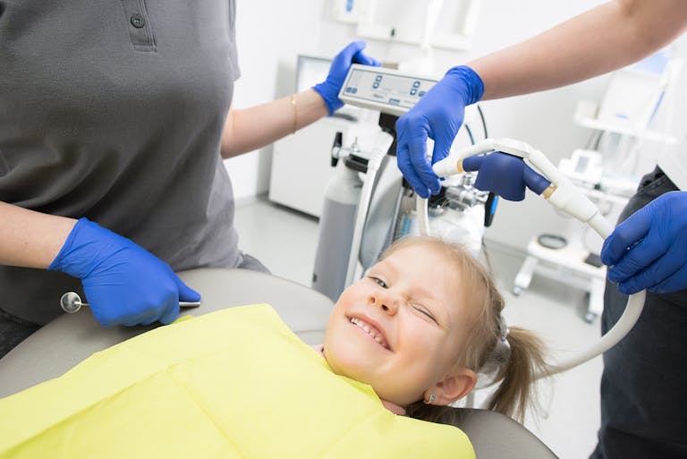 Child winking in a dental chair during a pediatric dental visit with staff and equipment nearby.