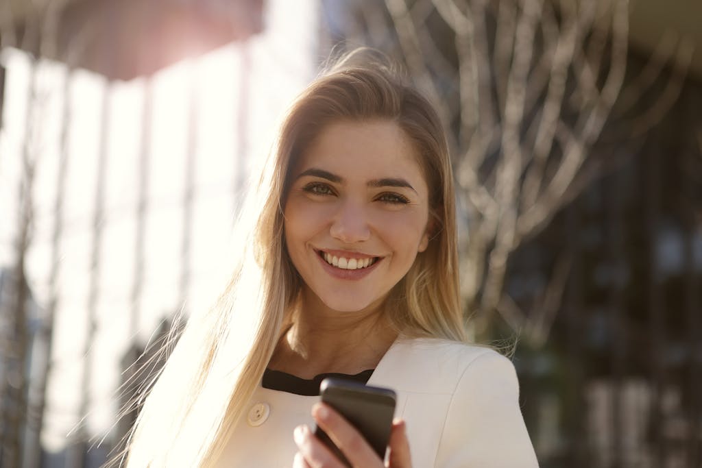 Smiling woman outdoors holding a smartphone in natural light.