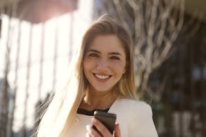 Smiling woman outdoors holding a smartphone in natural light.