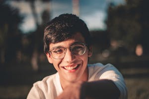 Young man with glasses smiling outdoors in warm sunlight.