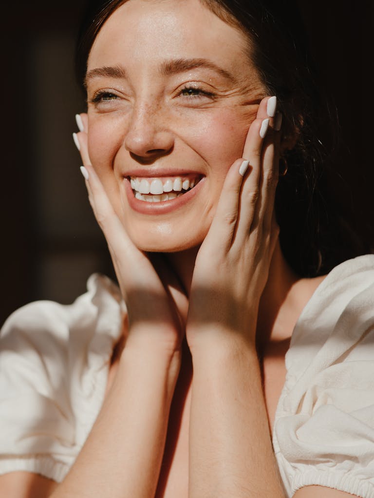 Smiling woman in natural light with hands on her cheeks, showing bright teeth.