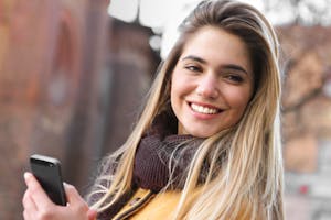 Young woman smiling outdoors while holding a smartphone and wearing a scarf and coat.