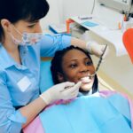 Dentist examining a patient’s teeth during a dental appointment while the patient holds a hand mirror.